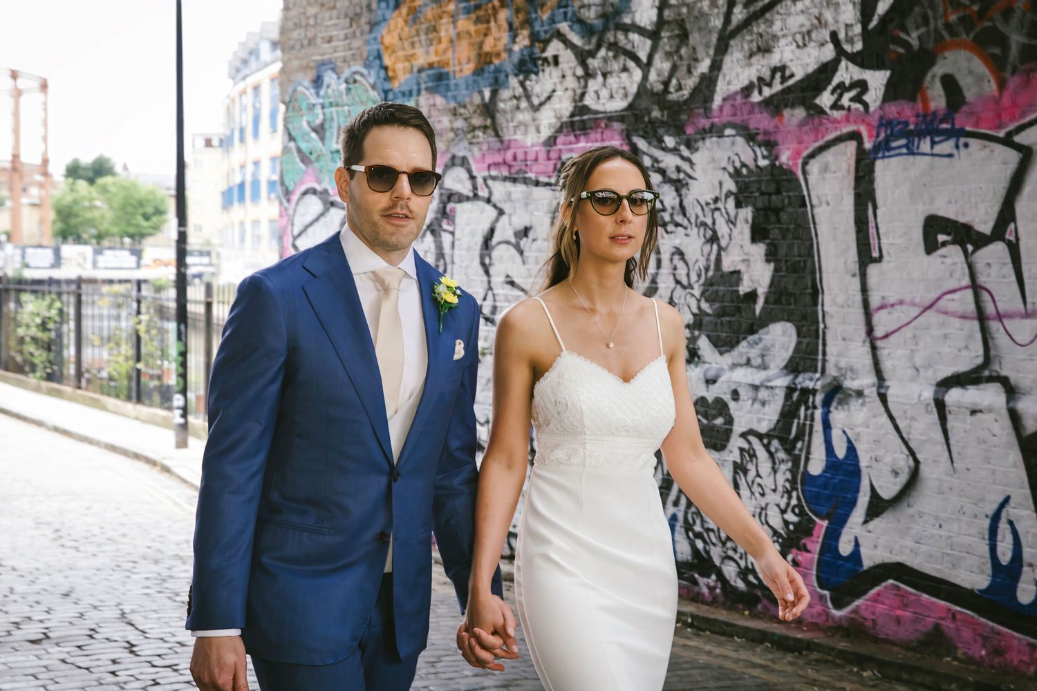 Bride and groom in sunglasses hold hands walking past colorful graffiti wall on cobblestone street.