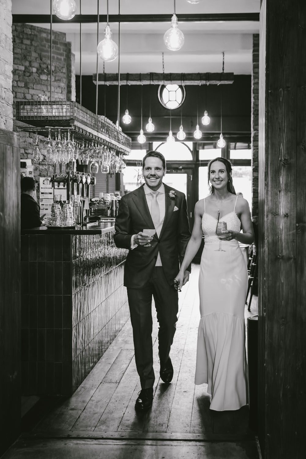 Black and white photo of a couple holding drinks and walking hand in hand through a stylish bar venue.