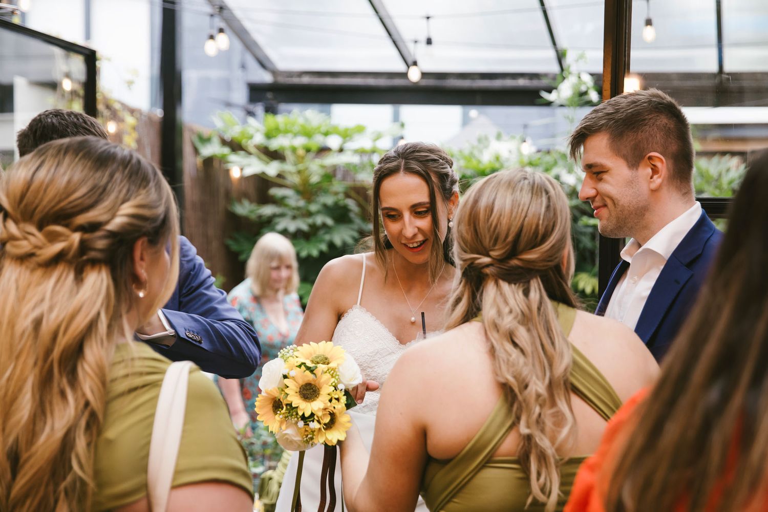 Bride holding sunflower bouquet surrounded by bridesmaids in olive dresses at a greenhouse wedding.