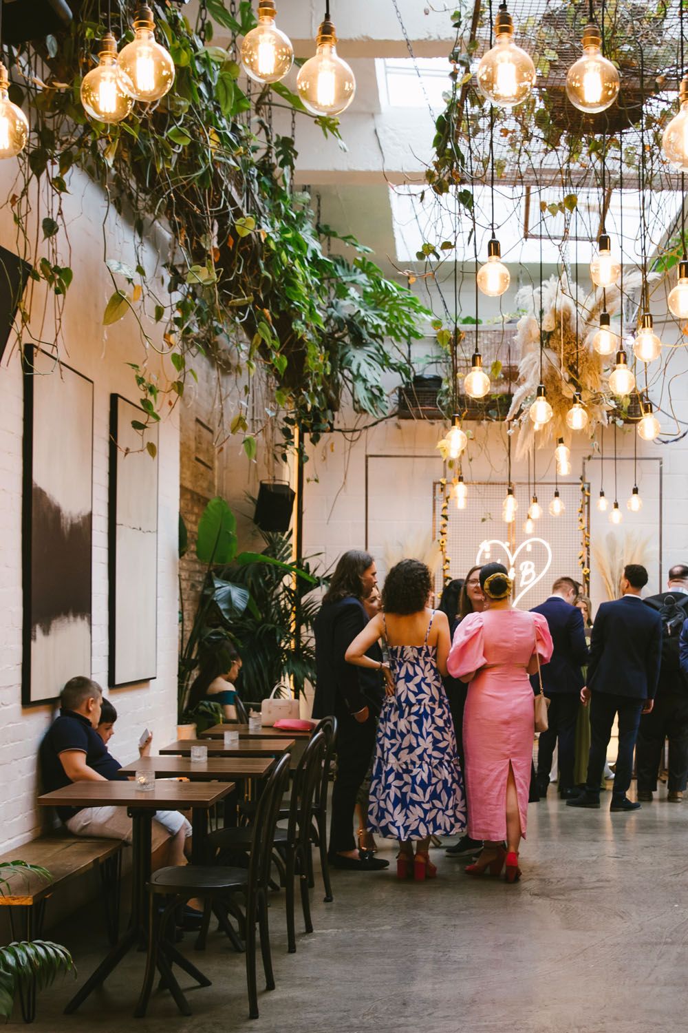 Trendy restaurant interior with hanging Edison bulbs, lush greenery, and elegantly dressed guests socializing.