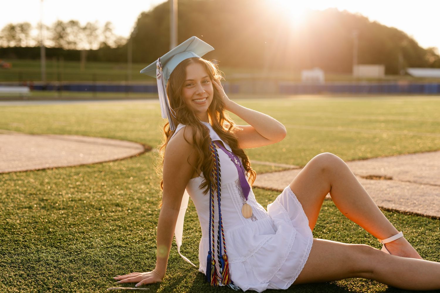 Smiling graduate in cap and gown sitting on field in golden sunset light.