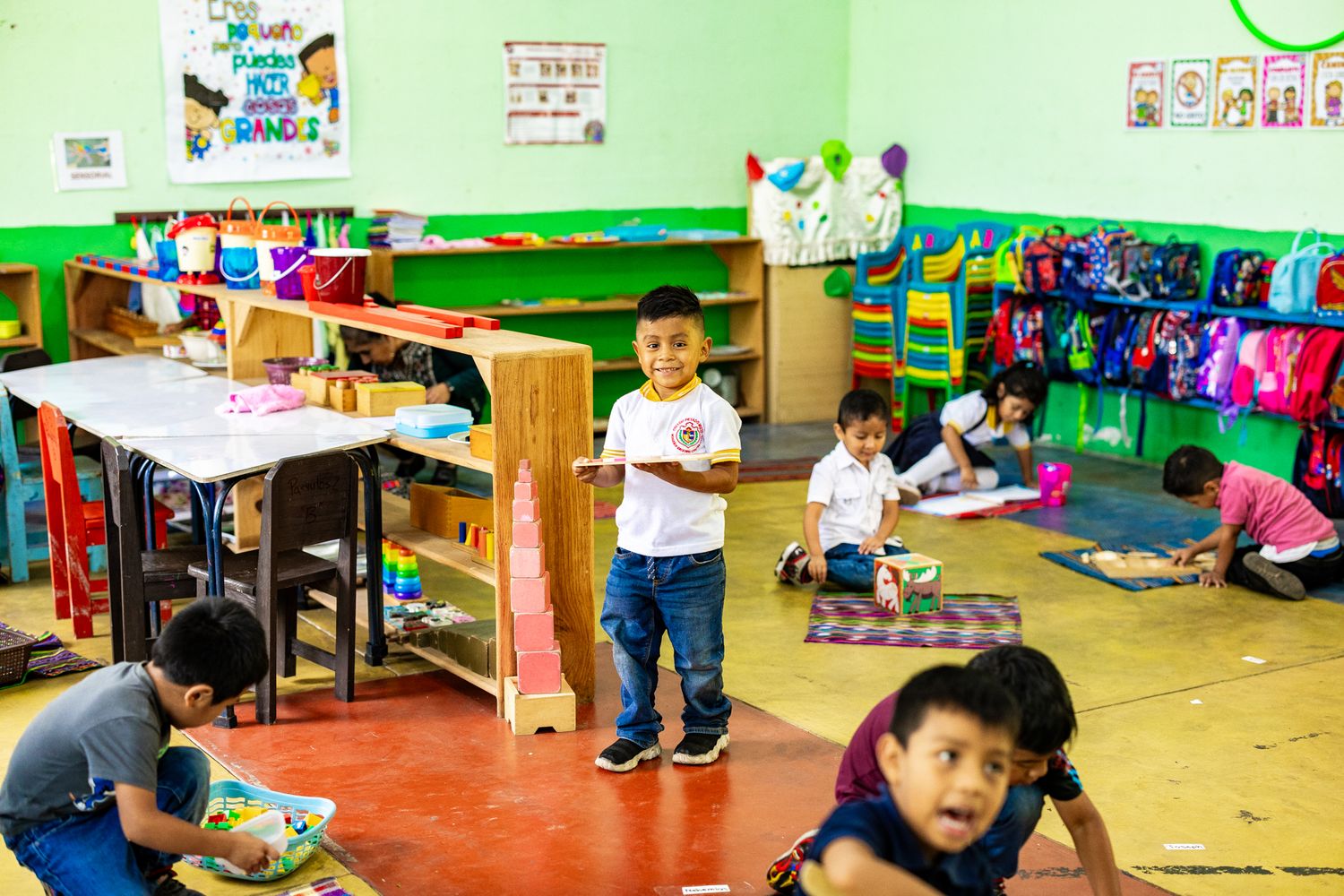 Children playing and learning in a colorful classroom with green walls, backpacks, and educational materials.