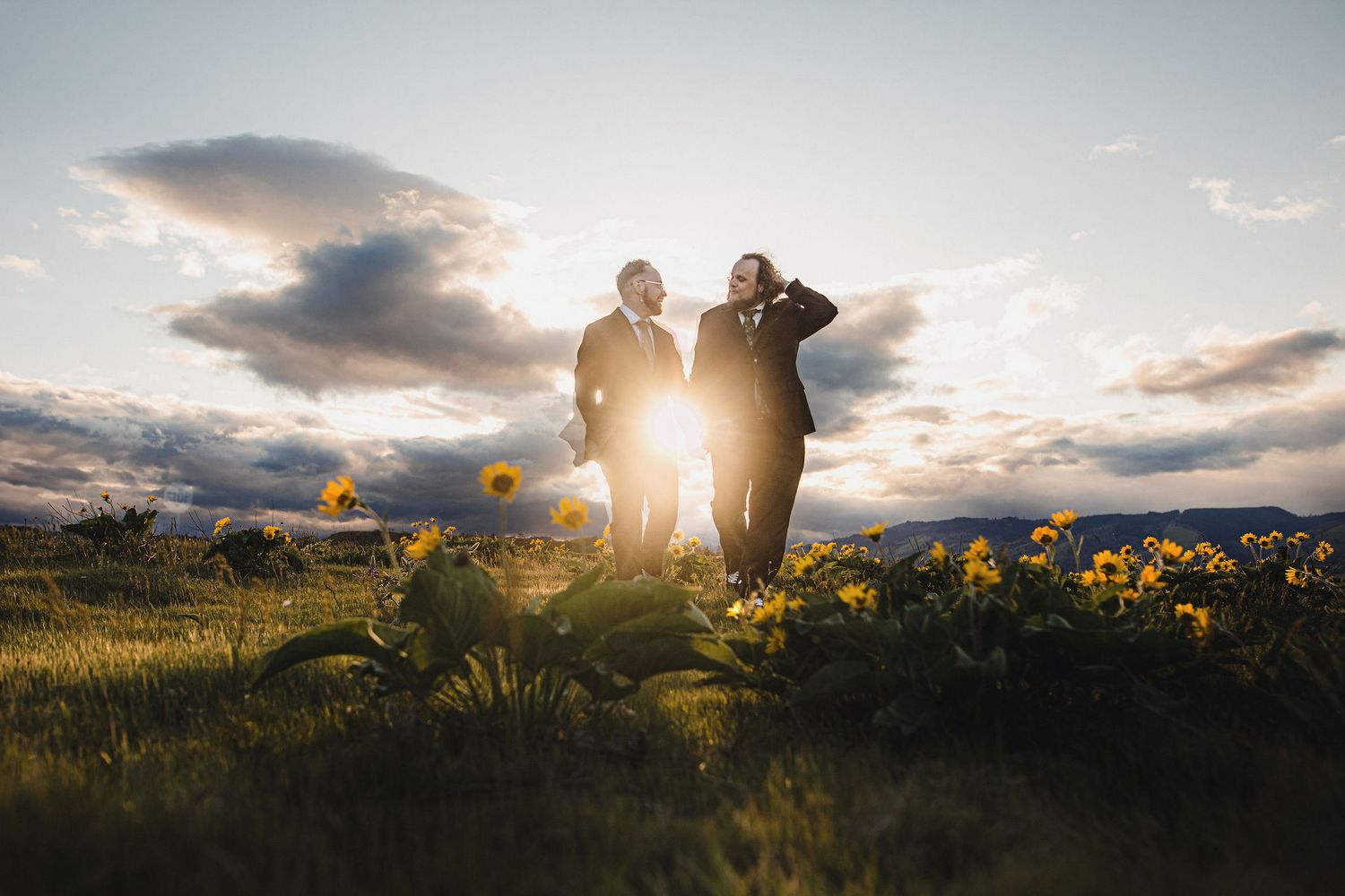 Two grooms stand in a sunflower field at golden hour, backlit by a stunning sunset with dramatic clouds.