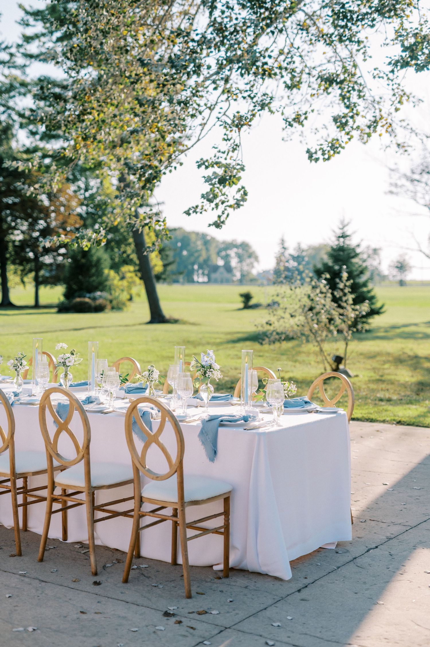 Elegant outdoor wedding reception table with gold chairs, white linens, and blue napkins set on a patio surrounded by lush greenery.