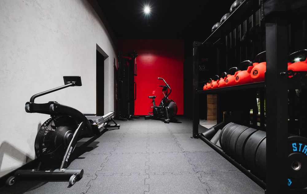 Nowoczesne wnętrze siłowni z ergometrami wioślarskimi, stojakami na ciężary, kettlebellami oraz czerwoną ścianą akcentową z rowerem treningo