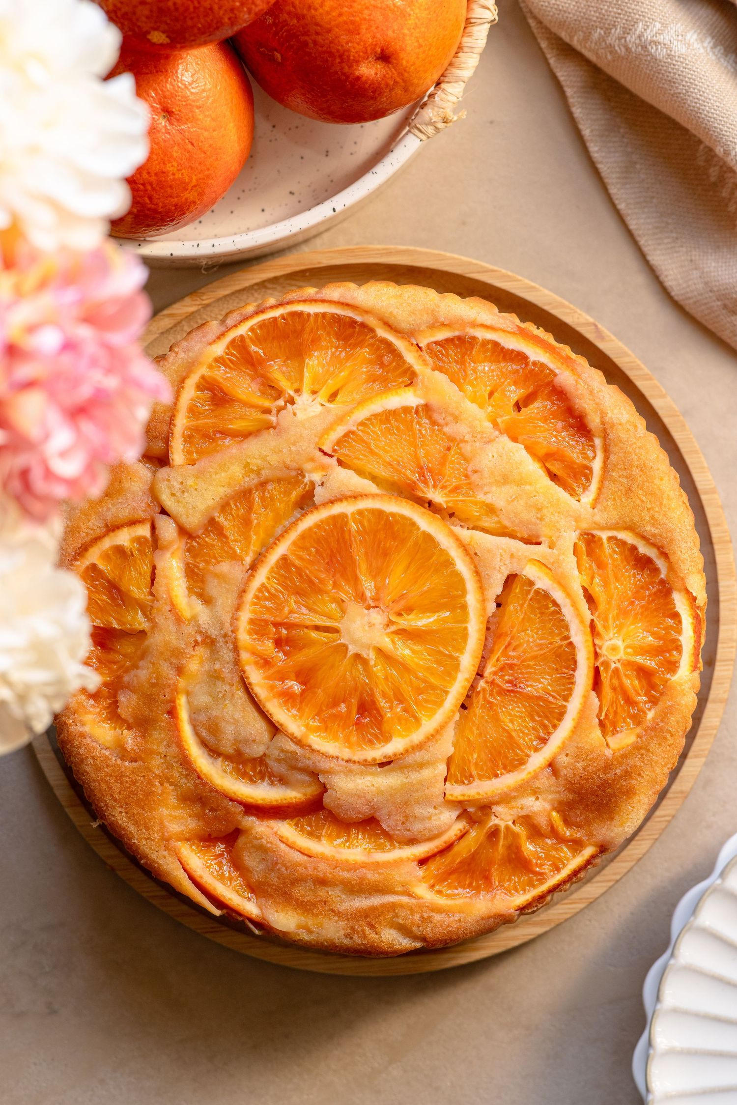 Upside-down orange cake topped with caramelized citrus slices on a wooden board, surrounded by fresh oranges and flowers.