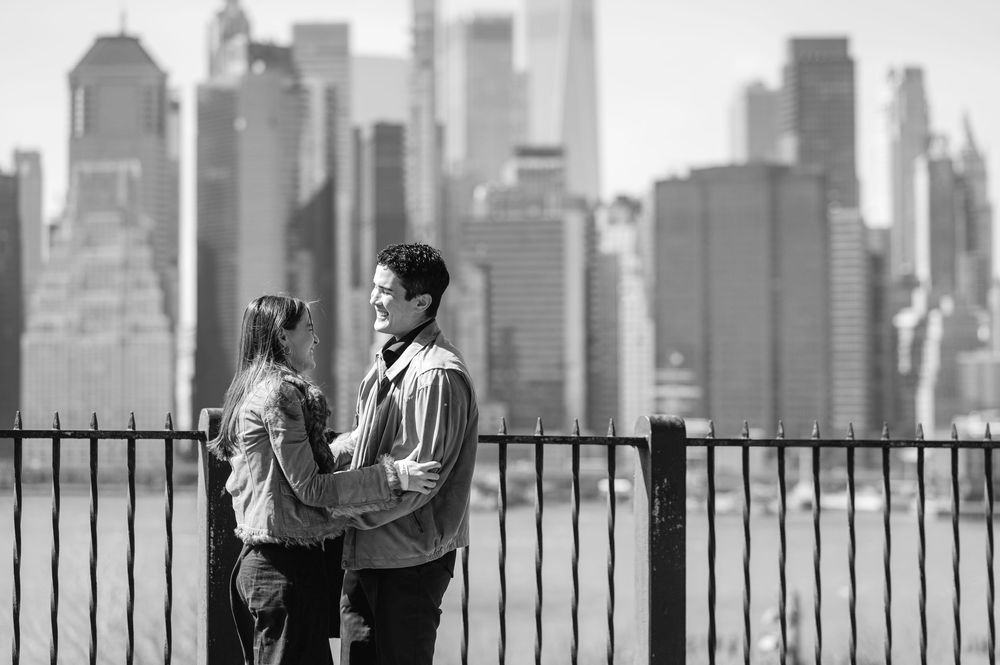 Black and white photo of couple embracing by iron fence with NYC skyline.