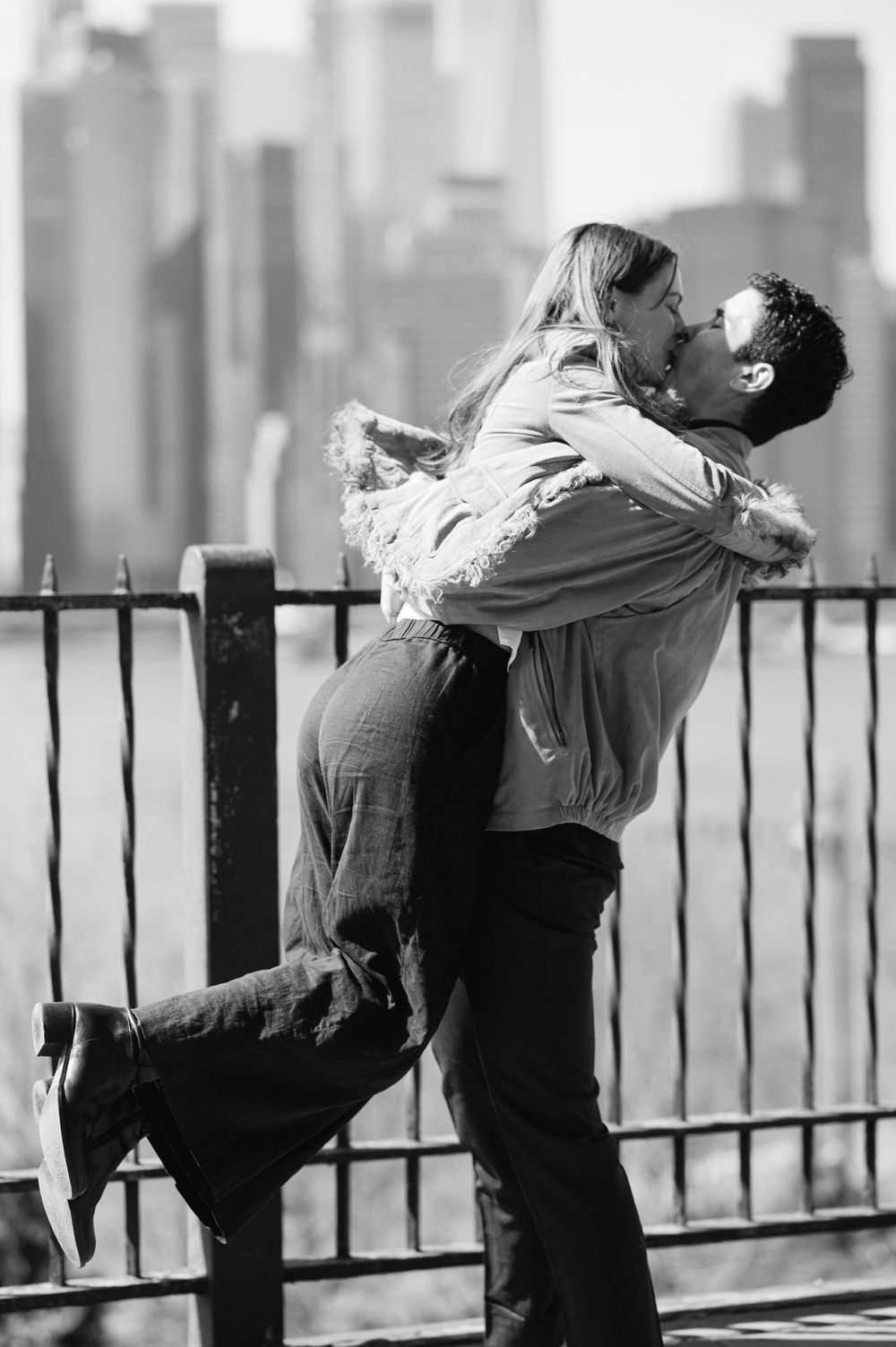 A man lifts and kisses a woman in a romantic black and white photo with a city skyline behind them.