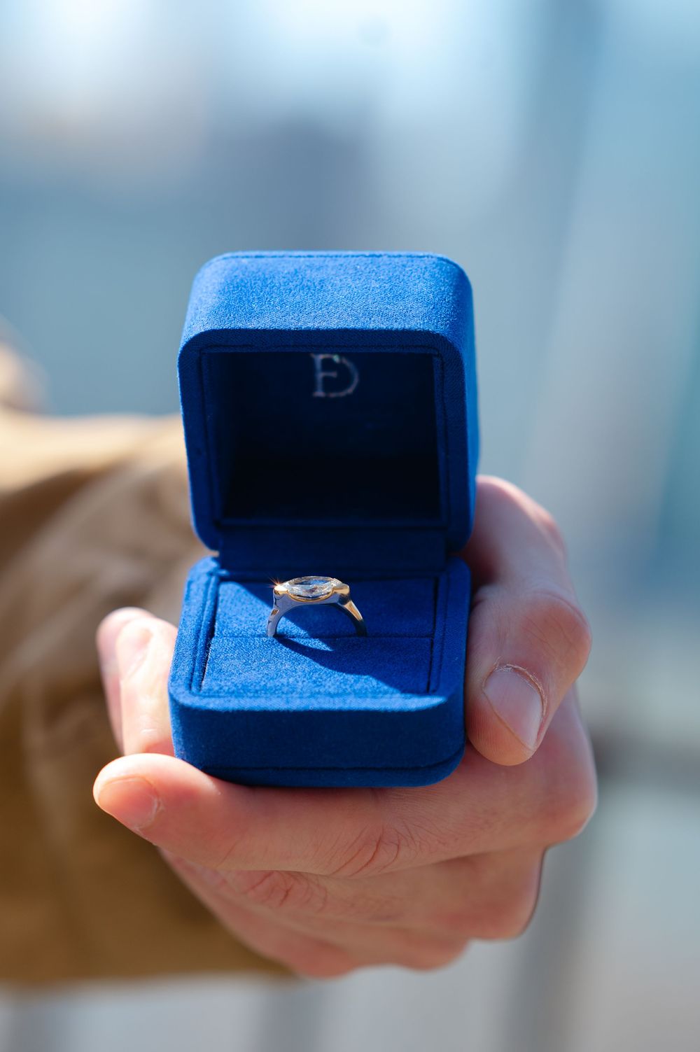 Hand holding open blue velvet ring box with diamond engagement ring inside.