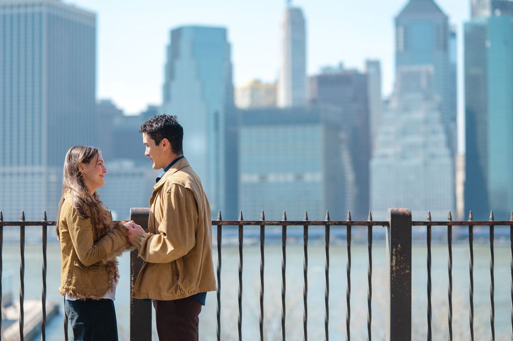 A couple in matching tan jackets stands by a railing with the New York City skyline in the background.