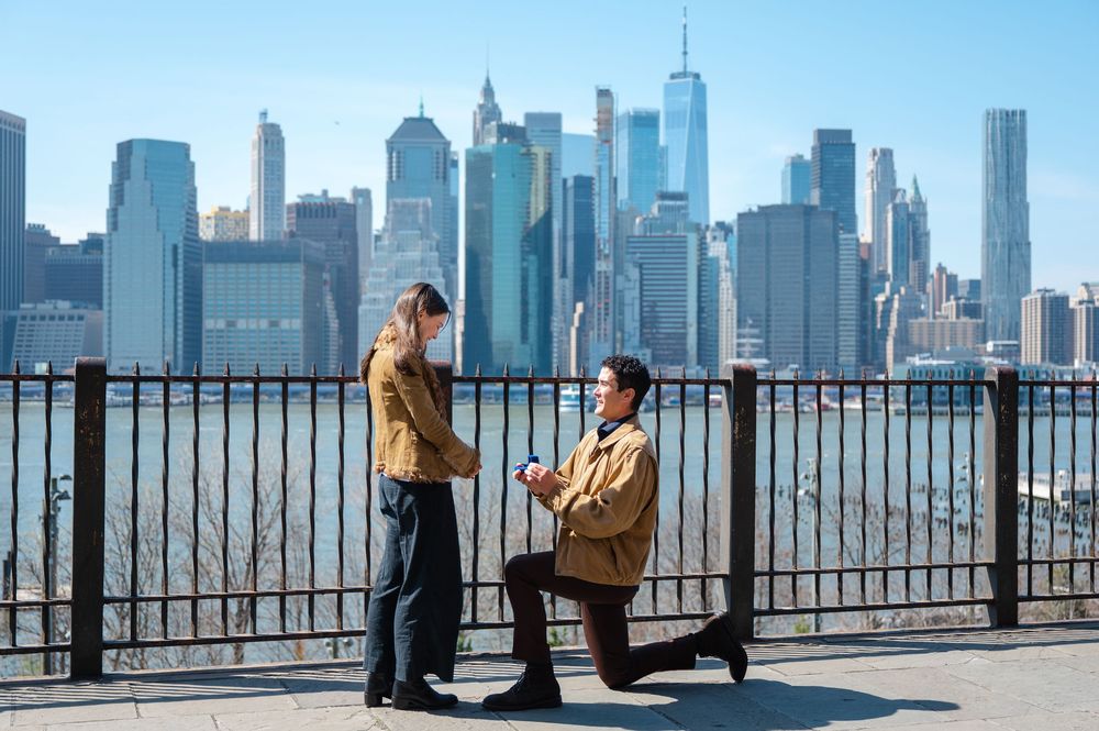 Man proposing to woman with NYC skyline backdrop at Brooklyn Bridge Park.