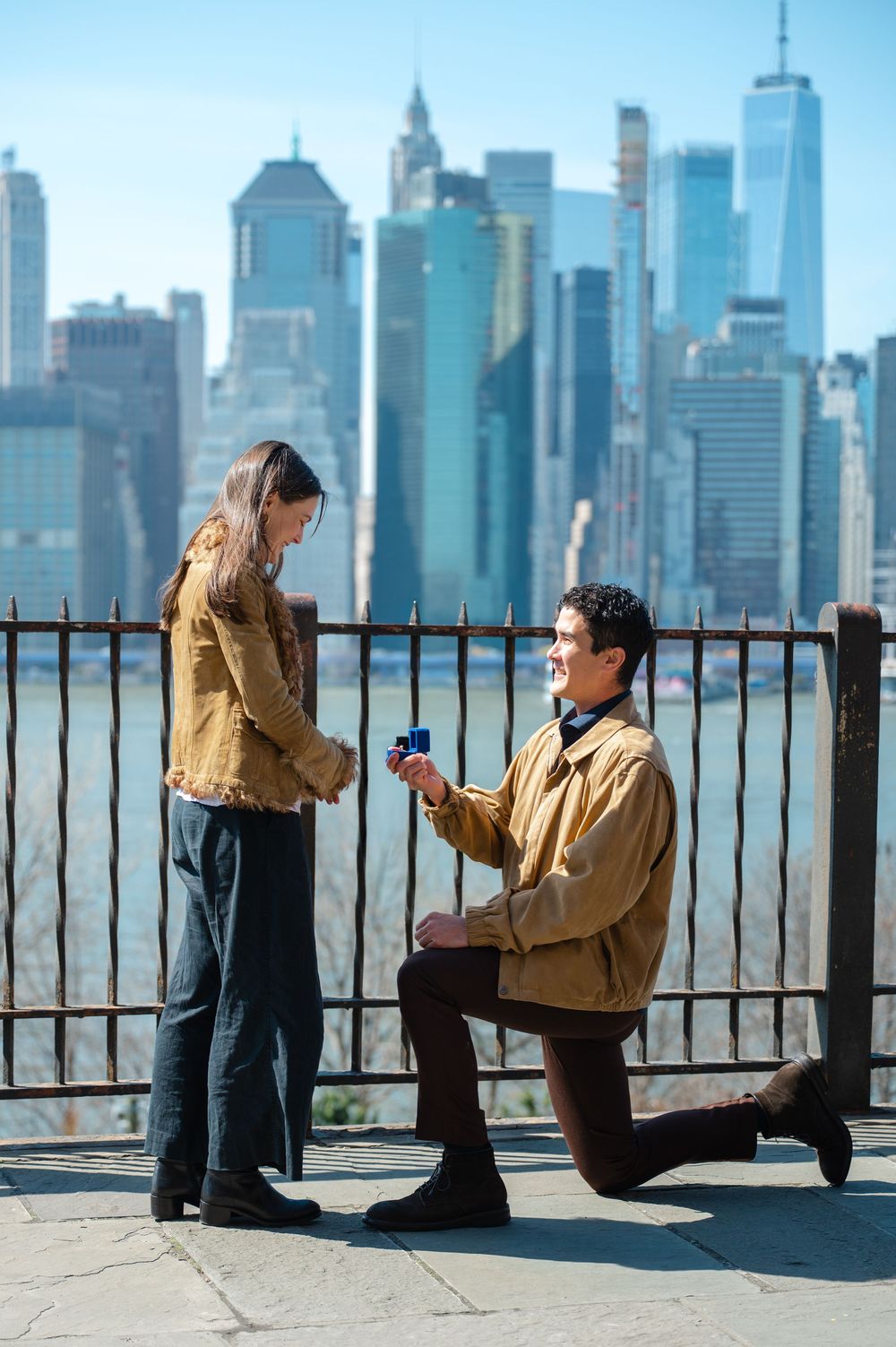 Man on one knee proposing to woman with NYC skyline backdrop on sunny day.