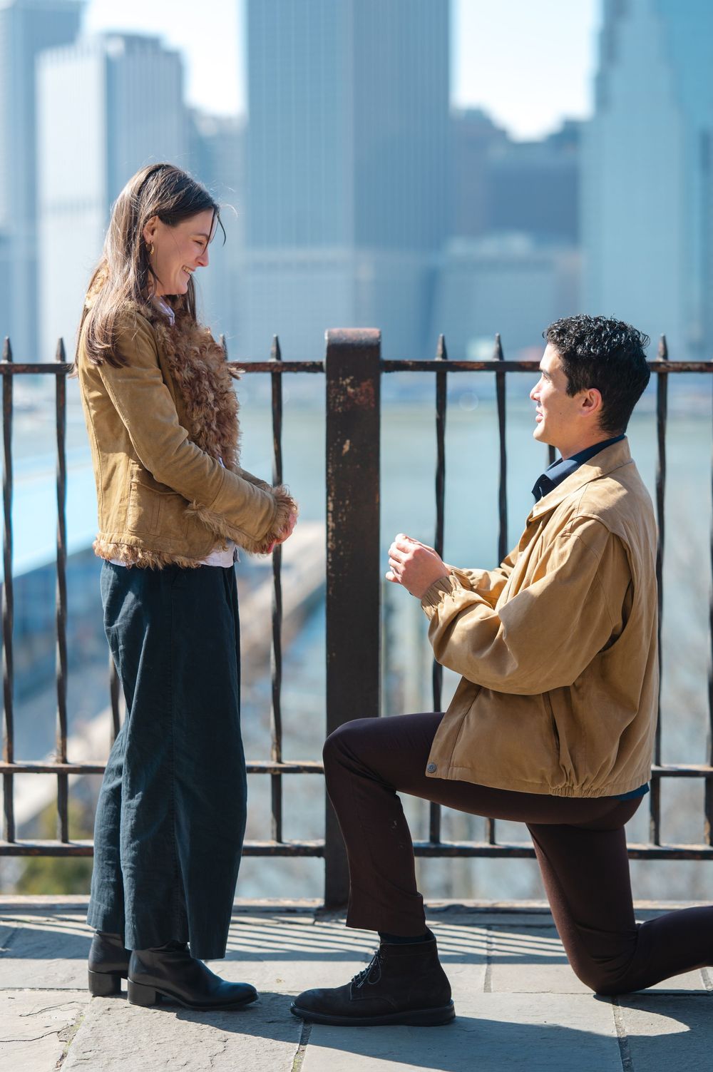 Man proposing on one knee to woman with NYC skyline backdrop.