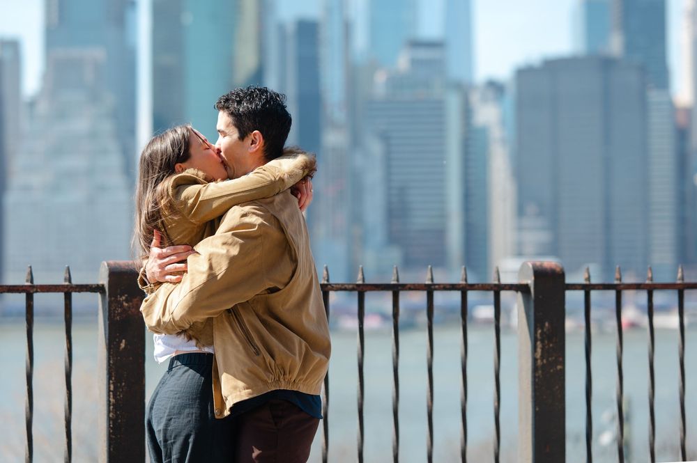 Couple embracing and kissing in front of NYC skyline by iron fence on sunny day.