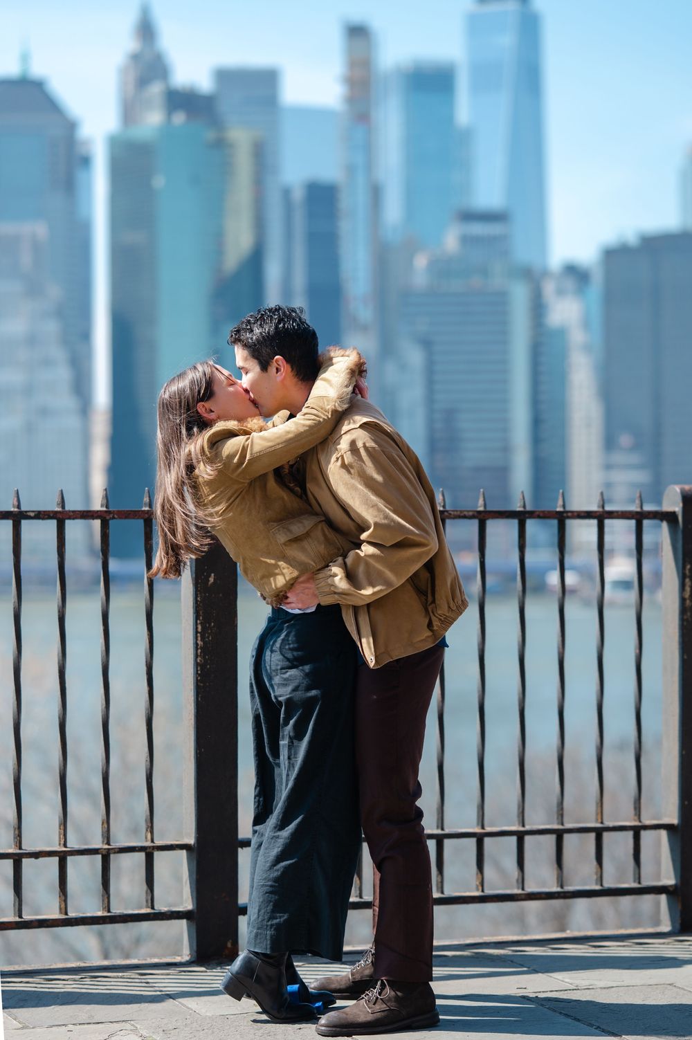 Couple kissing in front of a metal fence with the New York City skyline in the background.