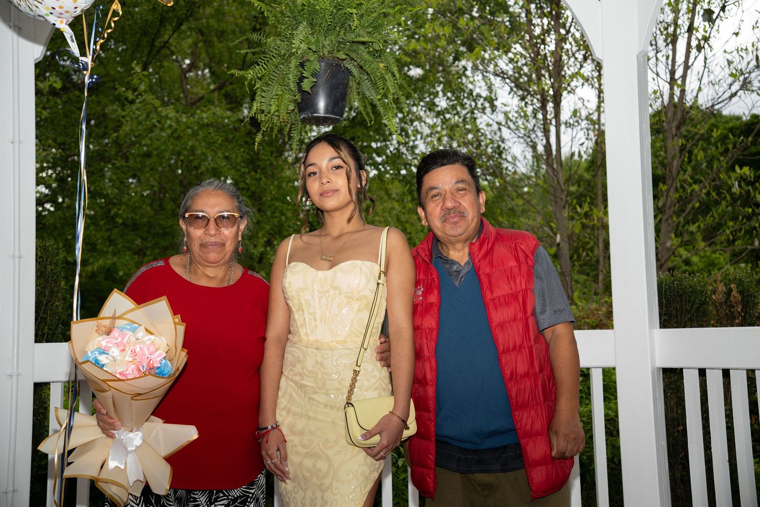 A young woman in a gold dress poses with two older adults holding a bouquet at an outdoor gazebo.