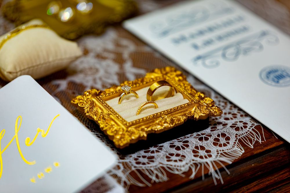 Gold wedding rings displayed in ornate frame with lace and wedding stationery on wooden table.