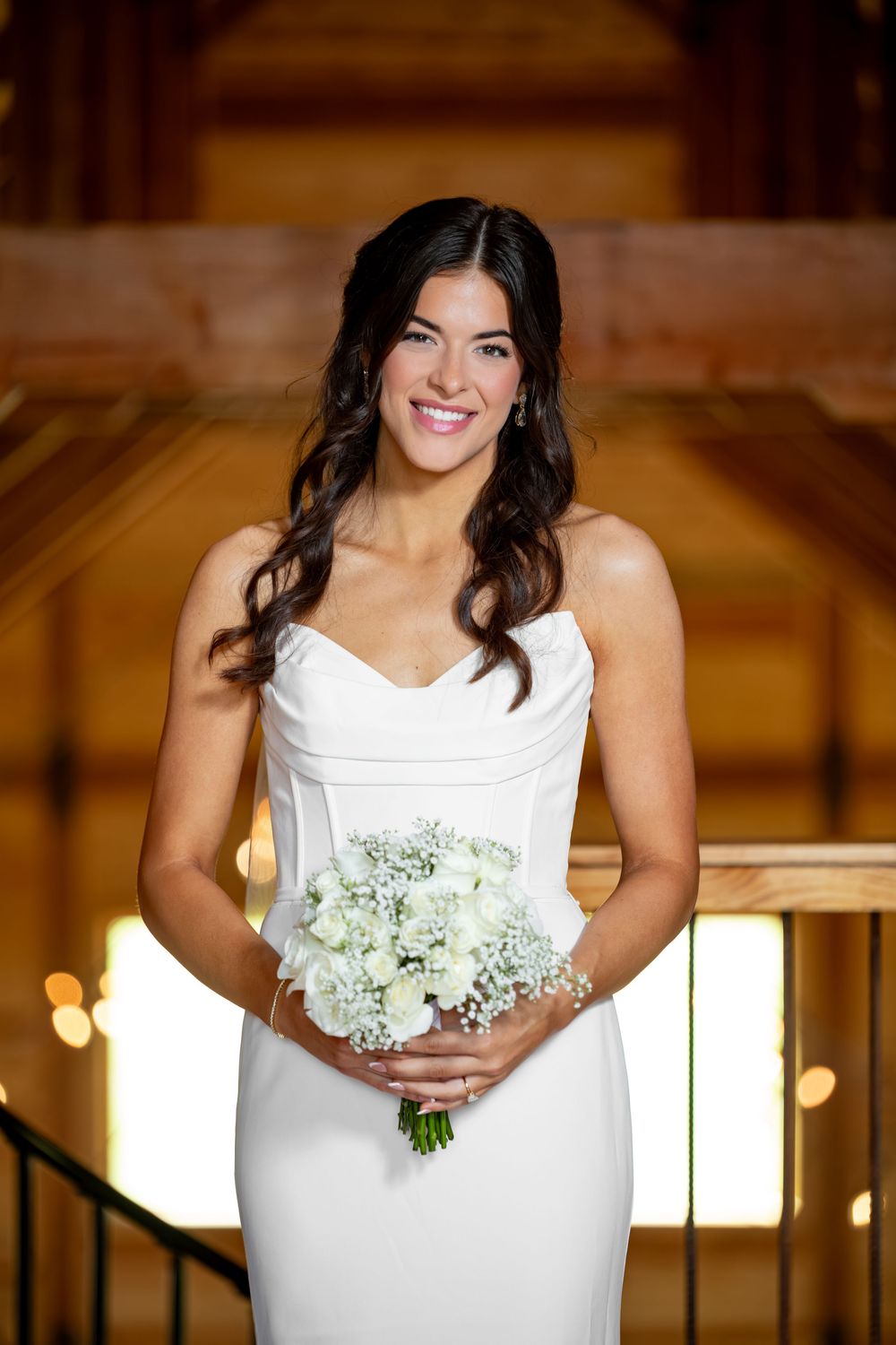 Smiling bride in white strapless gown holding white rose bouquet inside a warm wooden venue.