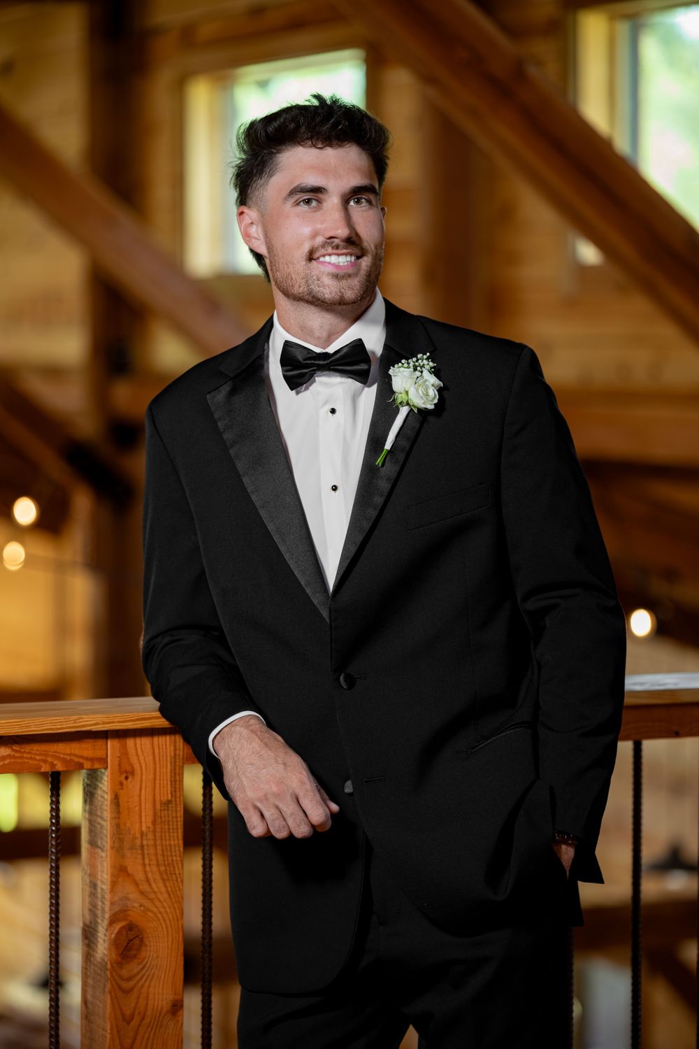 Groom in black tuxedo with boutonniere posing in rustic wooden venue.