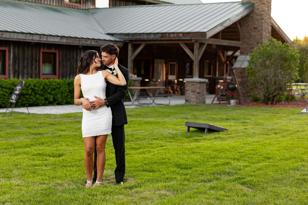 Couple in wedding attire embracing on green lawn outside rustic barn venue at dusk.