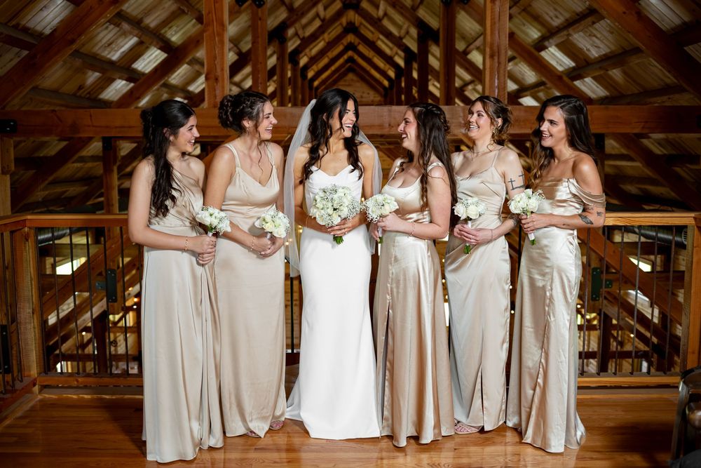 Bride in white gown stands with four bridesmaids in champagne satin dresses holding white bouquets in a rustic barn venue.