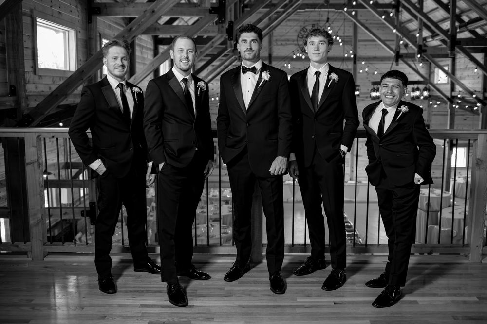 Groom and groomsmen pose in black suits on a rustic barn balcony in a classic black and white wedding photo.