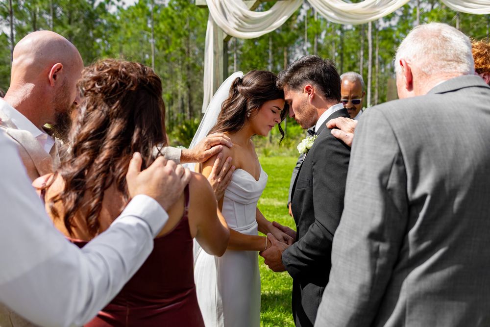 Outdoor wedding ceremony with bride and groom bowing in prayer, surrounded by guests placing hands on their shoulders.