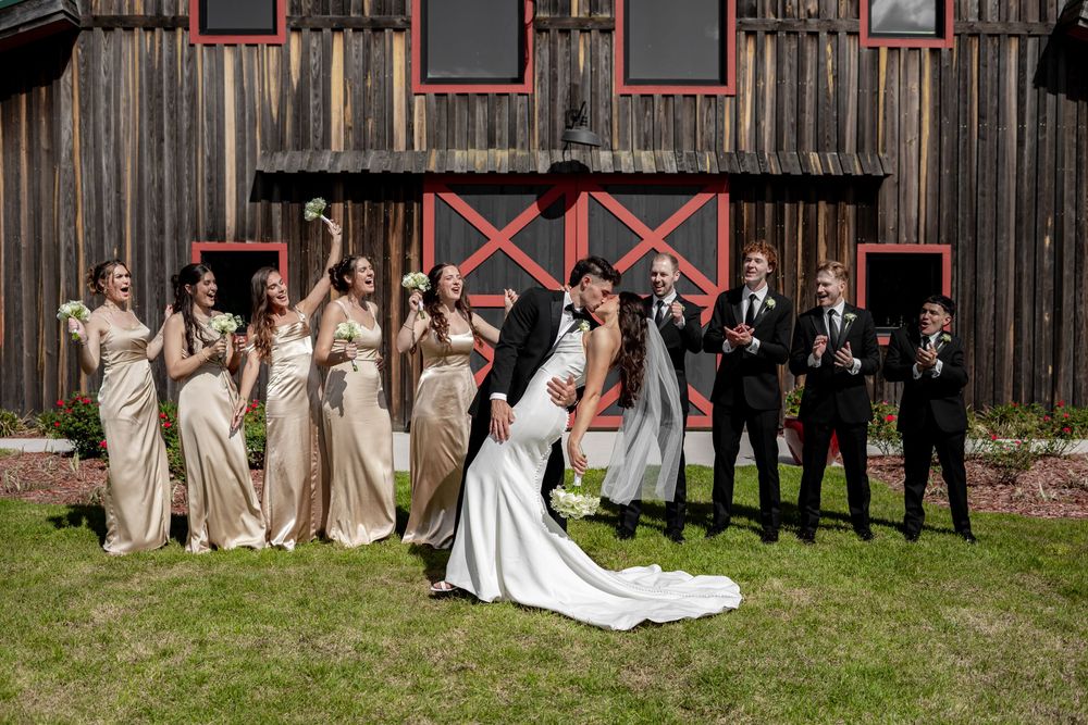 Bride and groom share a romantic dip kiss in front of a rustic red barn, surrounded by bridesmaids and groomsmen.