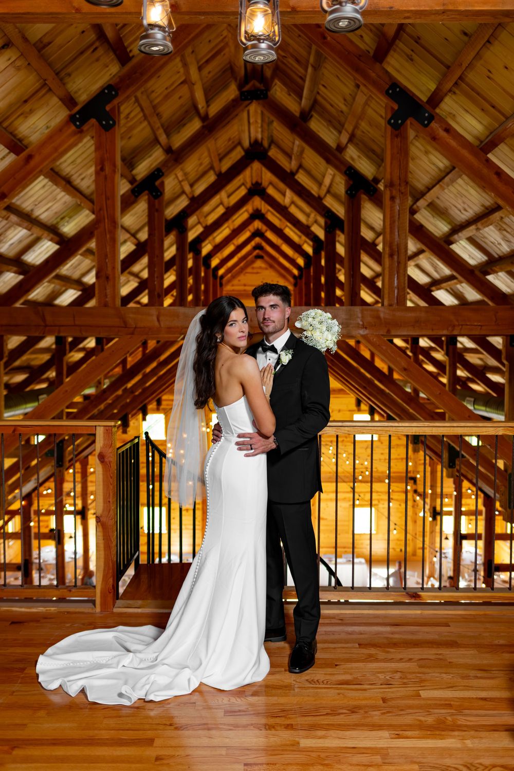 Bride and groom pose elegantly in rustic barn venue with wooden beamed ceiling and warm lighting.