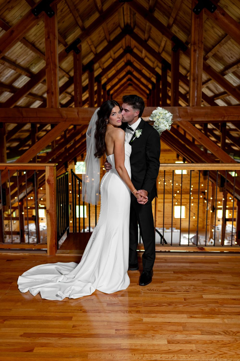 Bride and groom embracing in elegant barn venue, bride in fitted white gown with veil, groom in tuxedo.