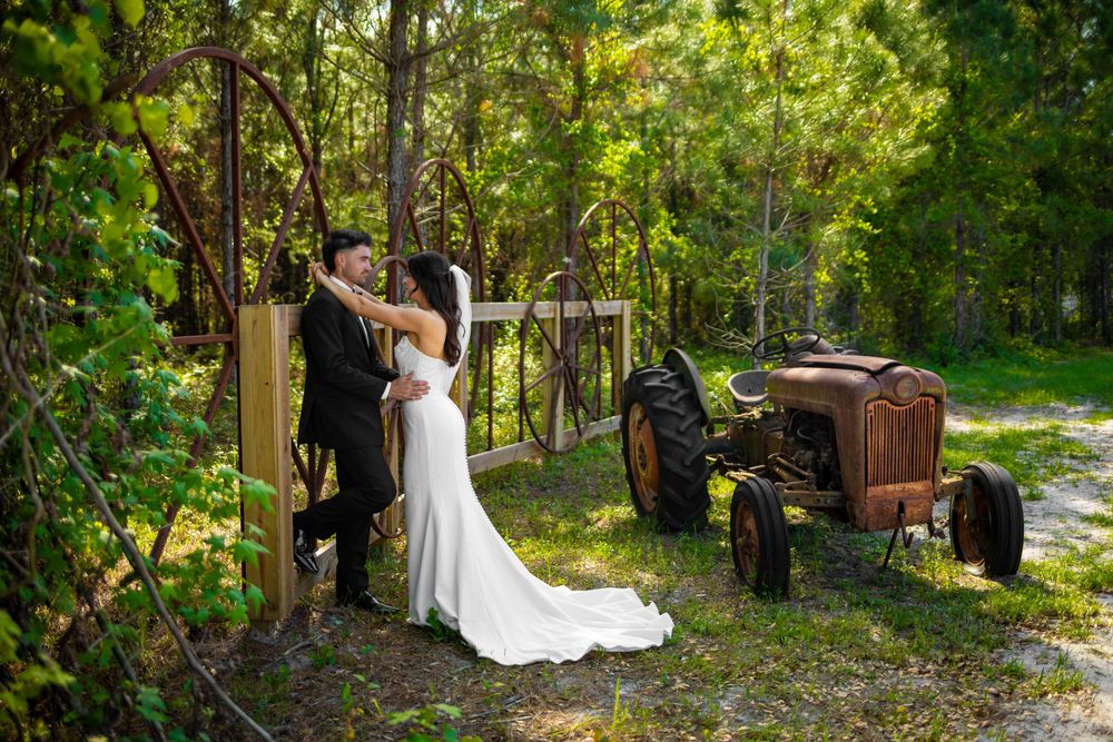Bride and groom share a romantic kiss near a rustic gate and vintage tractor surrounded by lush greenery.