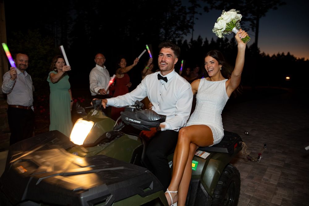 Bride and groom celebrate their wedding night riding an ATV outdoors while guests cheer with light-up wands.