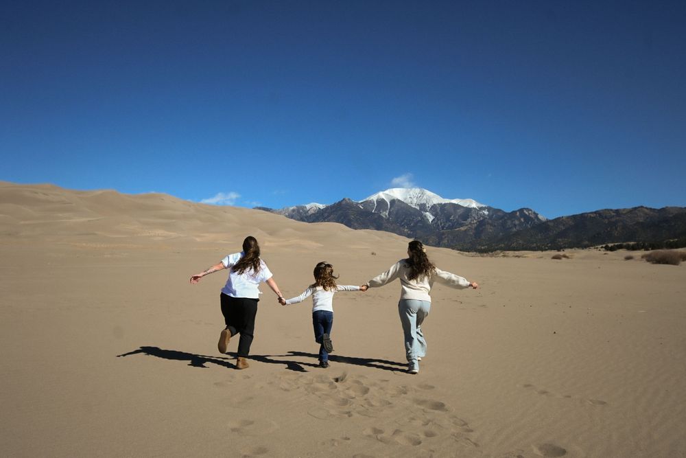 Three women hold hands running across sandy desert dunes with snow-capped mountains under a clear blue sky.