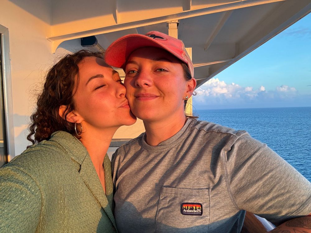 Two young women take a selfie on a cruise ship balcony at sunset with ocean views behind them.