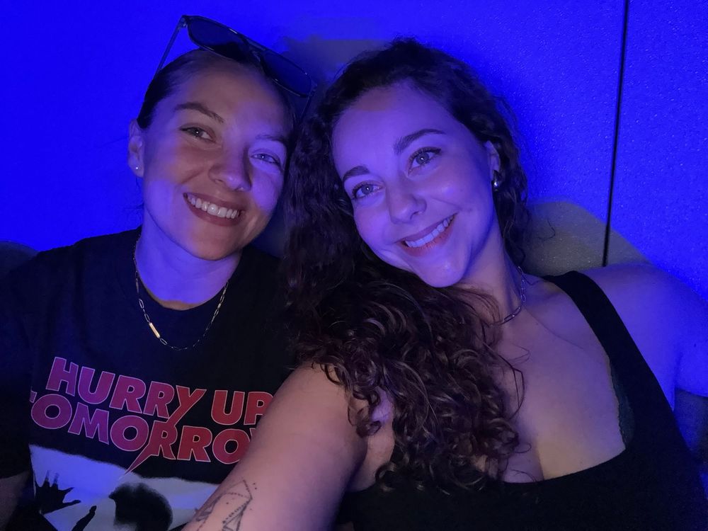 Two smiling women pose together under blue lighting, one wearing a black Hurry Up Tomorrow graphic tee.