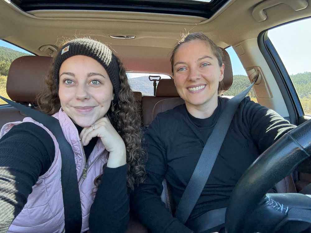 Two smiling women take a selfie inside a car on a scenic mountain road trip on a sunny day.