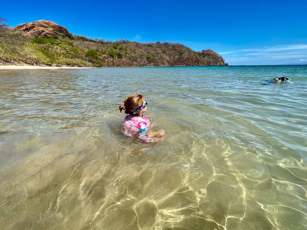 Young child sitting in shallow clear ocean water on a sunny tropical beach with green hills in the background.