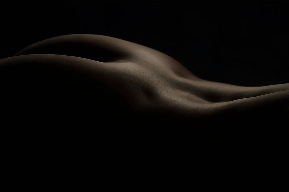 Studio-lit fine art nude study of a woman’s buttock and lower back in deep shadow.