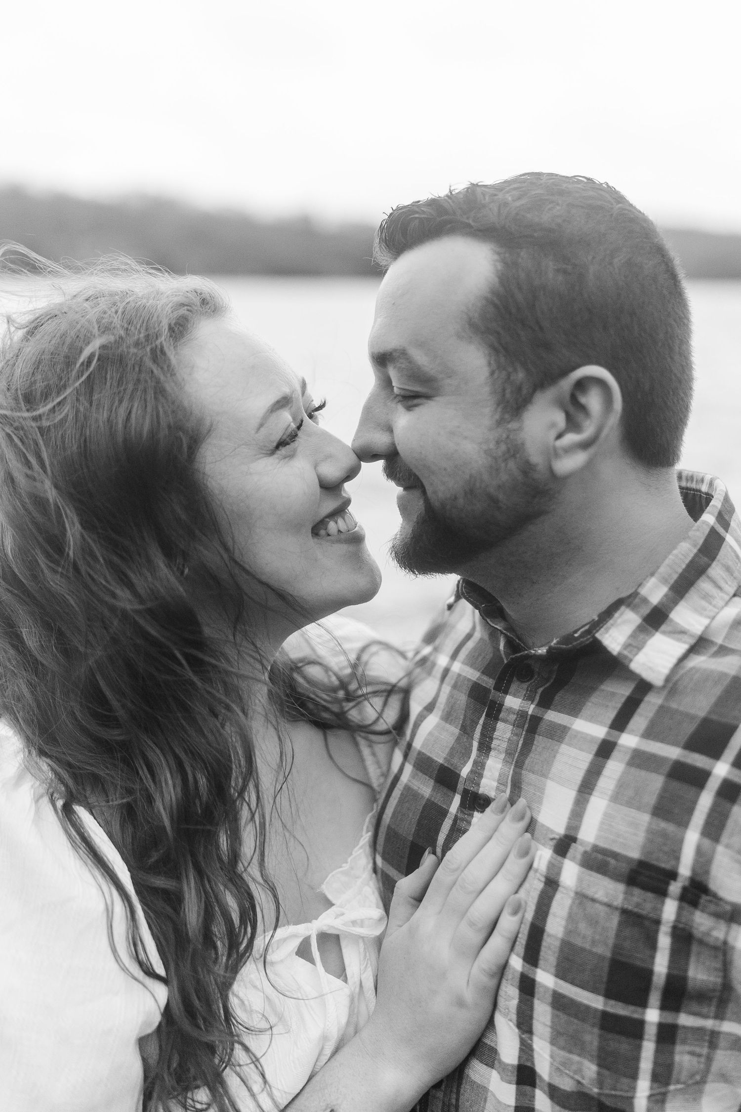 Black and white photo of a smiling couple about to kiss outdoors near a lake, woman with curly hair, man in plaid shirt.