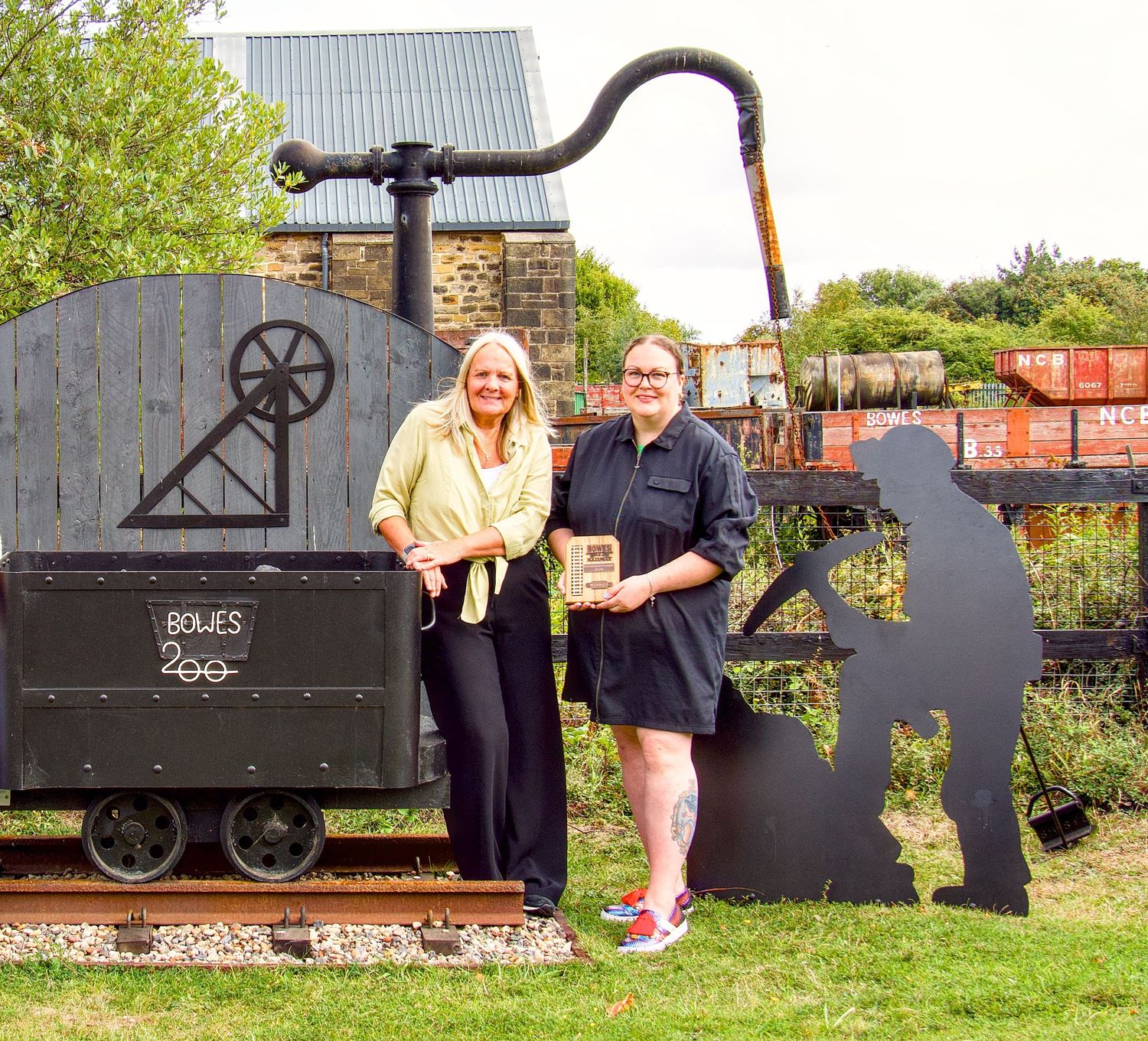 Two women posing beside a vintage mining cart at Bowes Museum outdoor heritage display on a sunny day.