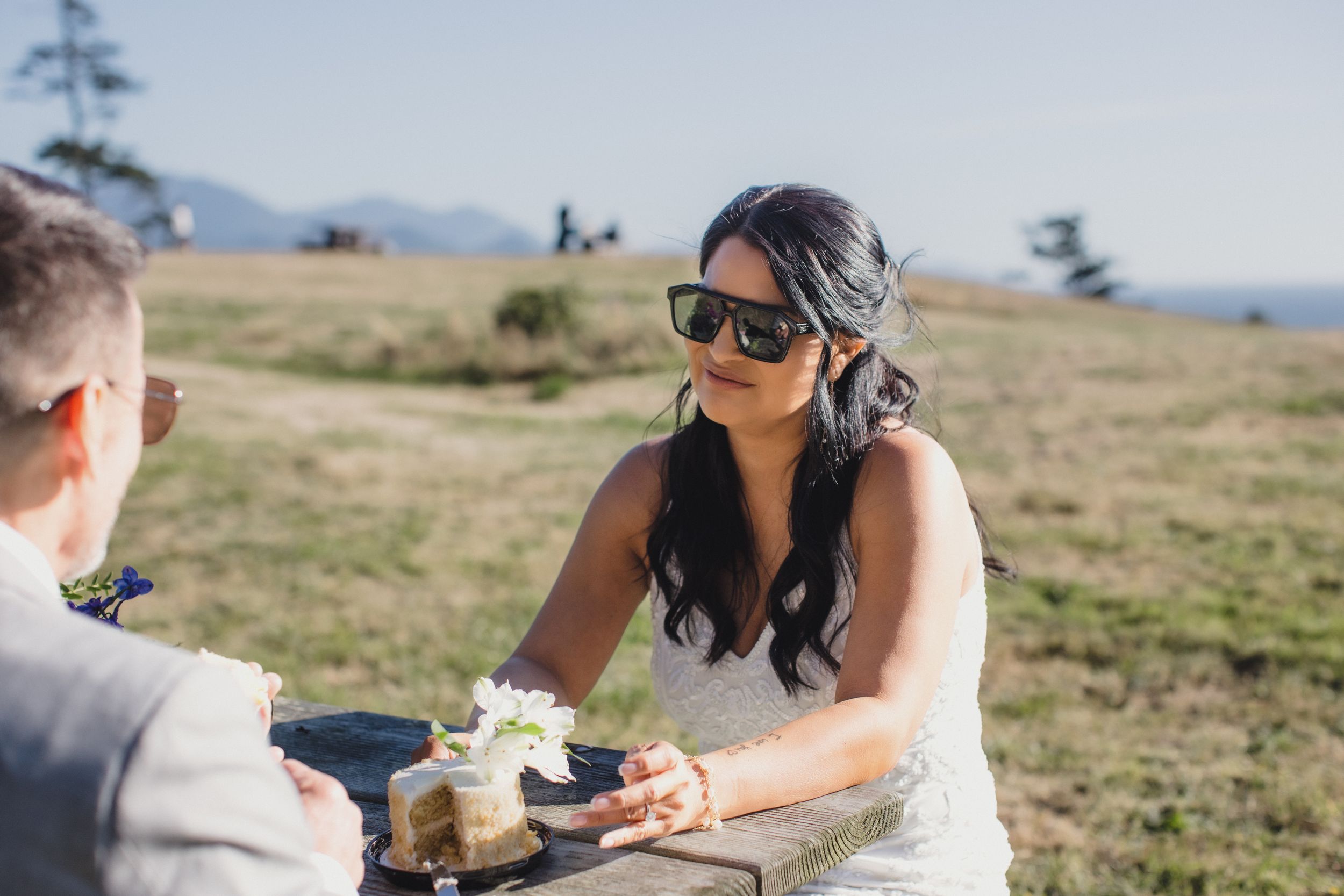 Bride in sunglasses sitting at outdoor table during wedding ceremony with scenic mountain backdrop.