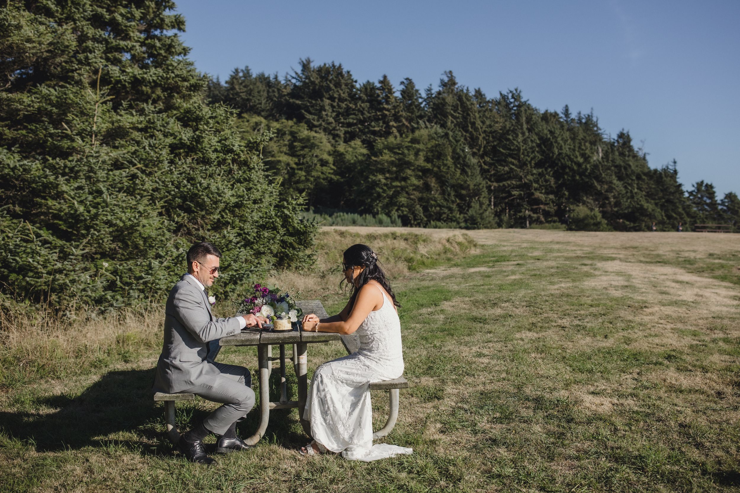 Bride and groom sitting at a rustic outdoor table in a green meadow with a forest backdrop on their wedding day.
