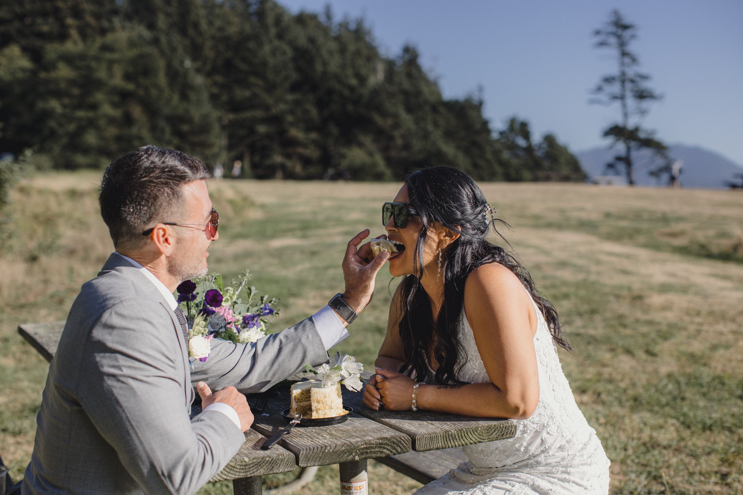 Groom in gray suit feeding cake to bride in white dress at outdoor picnic table with forest backdrop on wedding day.