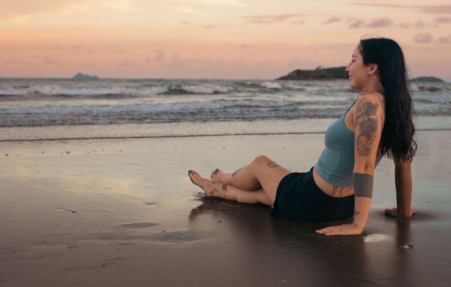 Woman with tattoo sitting on beach at sunset, waves in background, wearing crop top and skirt.