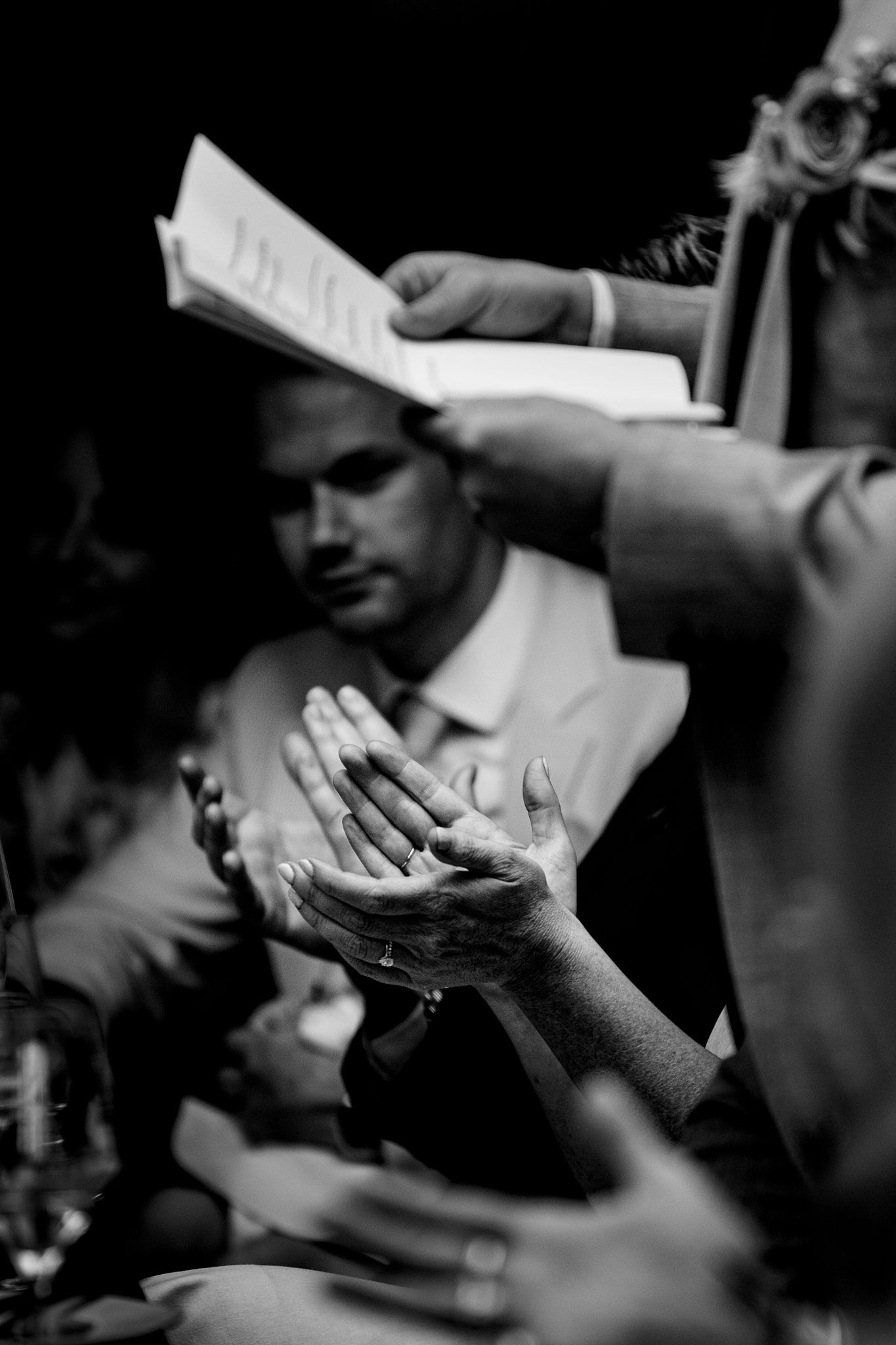 Black and white photo of wedding guests clapping hands at a reception dinner table, with a man in suit watching at Saraguay House