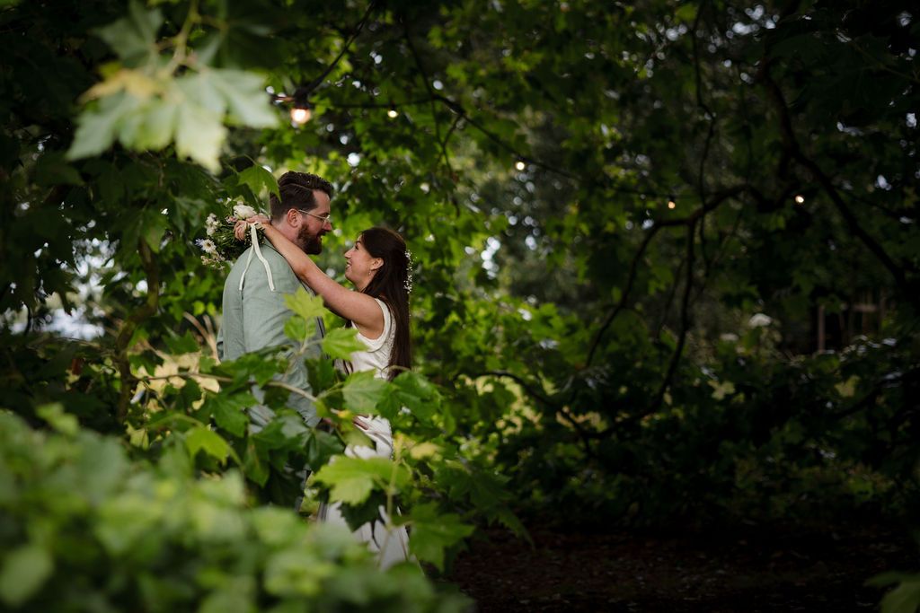 Couple embracing romantically among lush green leaves and trees in an outdoor garden setting.