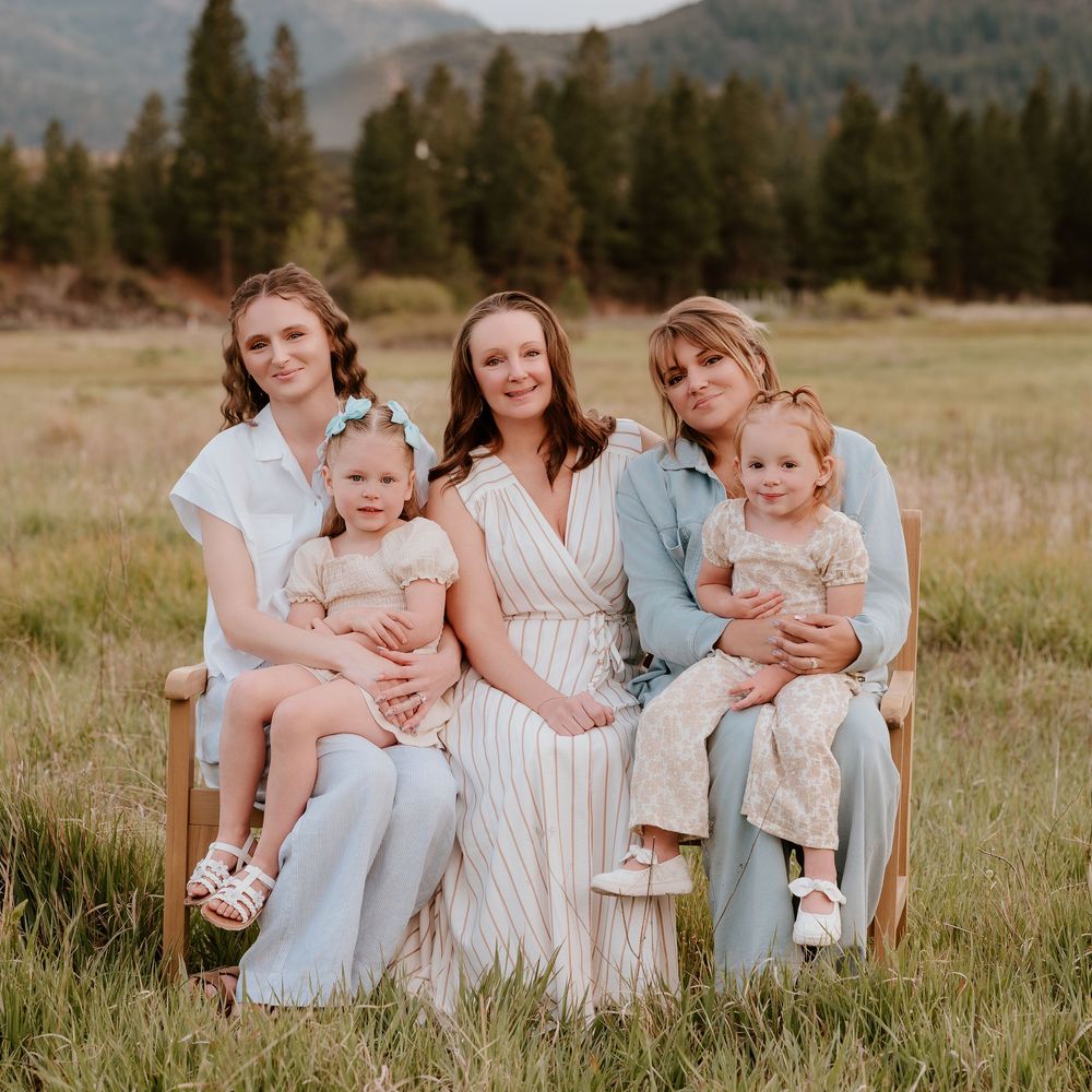 Three women and two toddler girls pose together in a scenic mountain meadow for a family portrait.