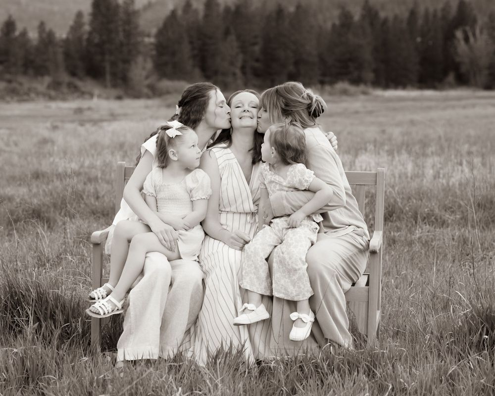 Black and white photo of a mother sitting in a field while her three young daughters kiss her cheeks outdoors.