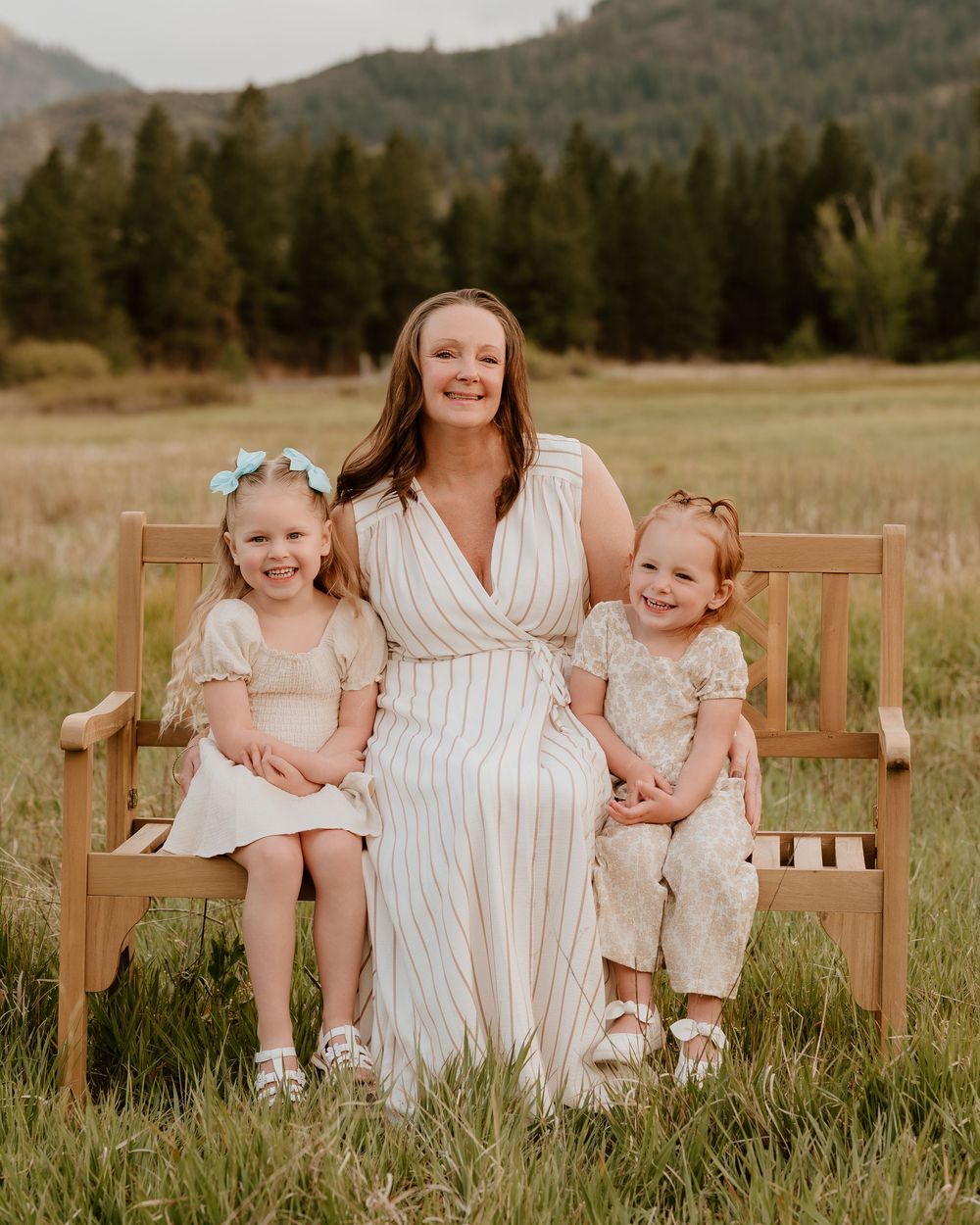 A woman in a striped dress sits on a wooden bench with two young girls in a scenic mountain meadow.