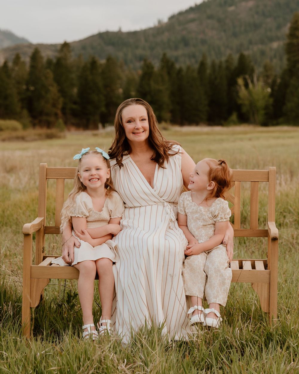 A woman in a striped dress sits on a wooden bench with two young girls in a meadow with mountains behind.